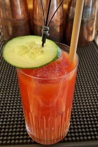 a rich red colored drink in a highball glass with an cucumber slice on top as a garnish