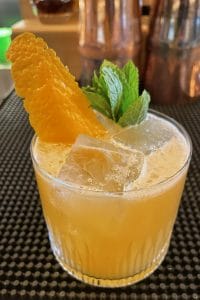 a shiny orange cocktail in a rocks glass with an orange peel and mint as a garnish